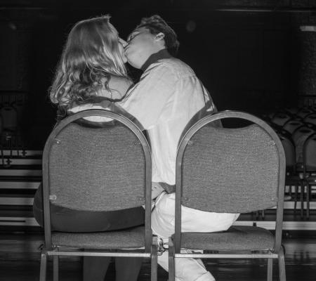 black and white photo of a pair kissing while seated with the chair backs turned toward the camera