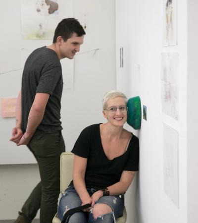 person sitting with ear pressed to small green sculptural object mounted on wall