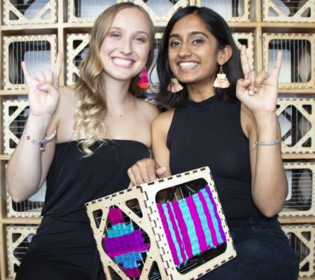 two women students holding up weaving project and their hands in a hook em gesture