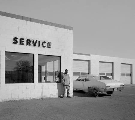 Black and white photograph of a man leaning against a service station wall taken by photographer Bryan Schutmaat