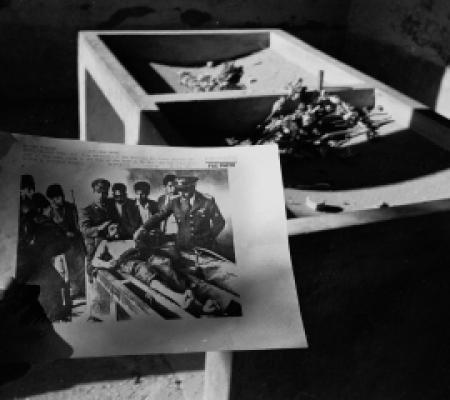 the photo is in black and white. a man is holding a photo of men in military uniforms gathered around a shirtless man in a stretcher. he seems hurt or sick.
