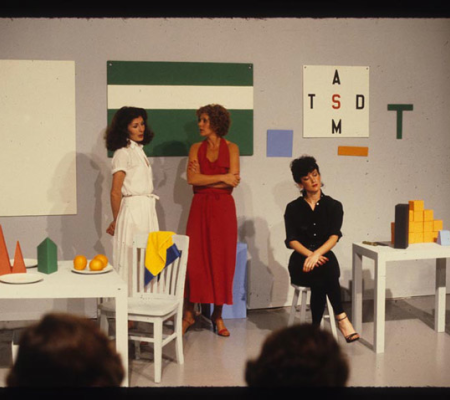 3 women on a stage surrounded by colorful, geometric objects and artworks on the wall, with audience members in foreground