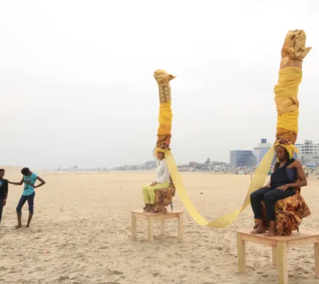 four women on beach, two standing and two seated on wooden platforms wearing sculptural head pieces 