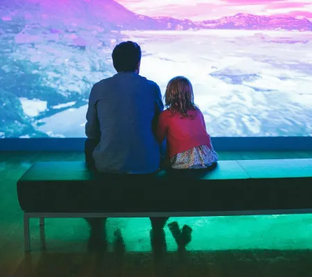 adult and child sitting on bench watching video installation