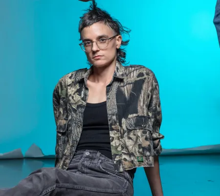 riel Sturchio sitting on the floor in a studio with a blue background. Their hair is being fixed by a disembodied hand (the hand of friend Dr. Kanwal Matharu). The photo was taken by Thalassa Raasch.